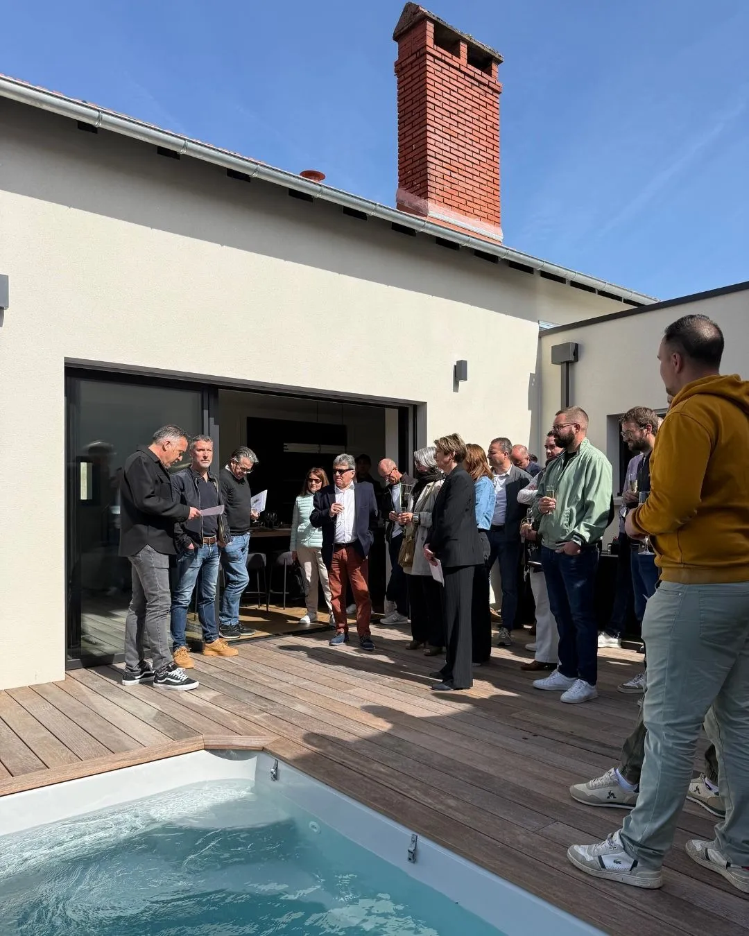 Visiteurs réunis sur la terrasse en bois d'une maison rénovée, autour d'un bassin, sous un ciel bleu.