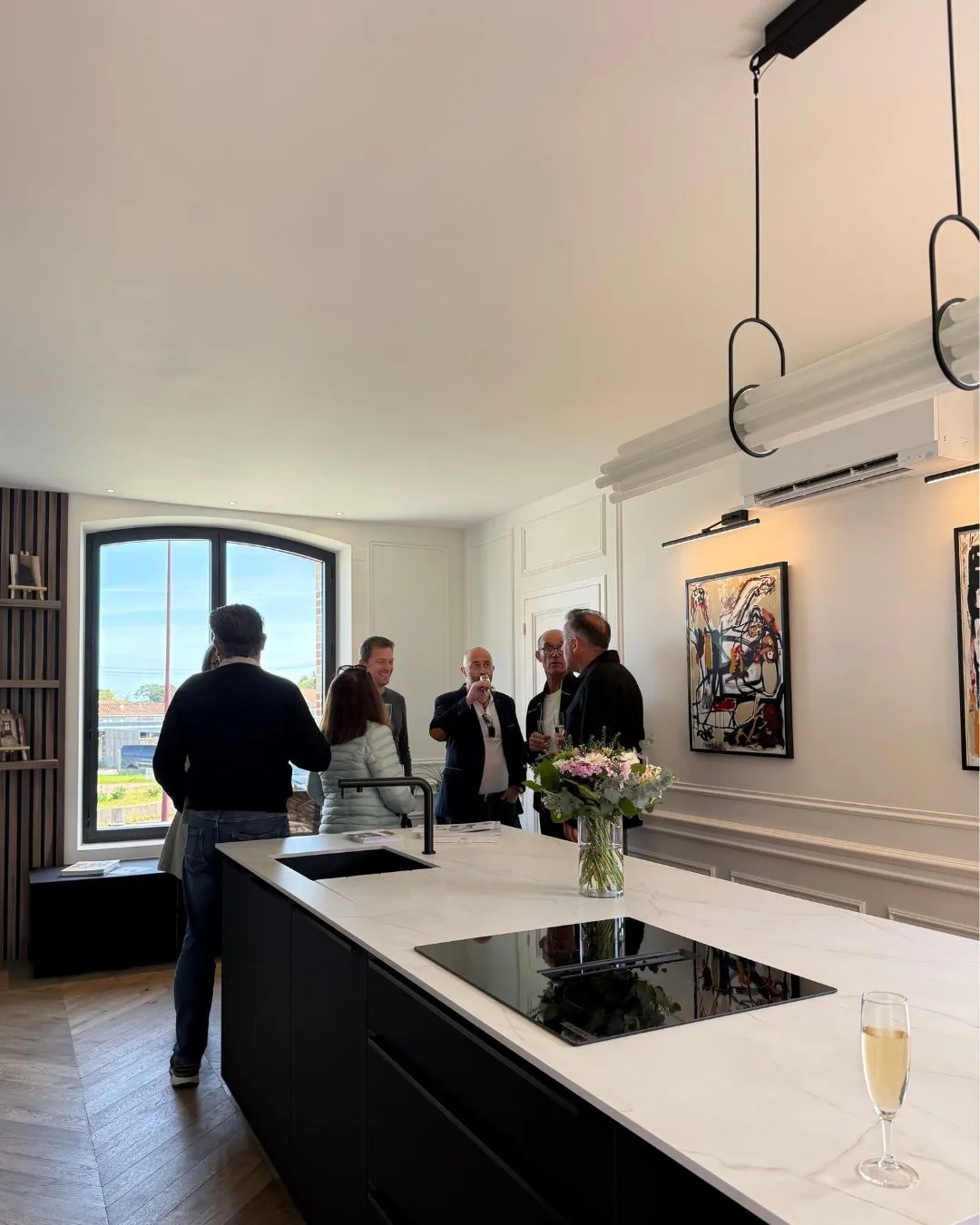 Visiteurs autour de l'îlot noir de la cuisine, une coupe de champagne au premier plan.