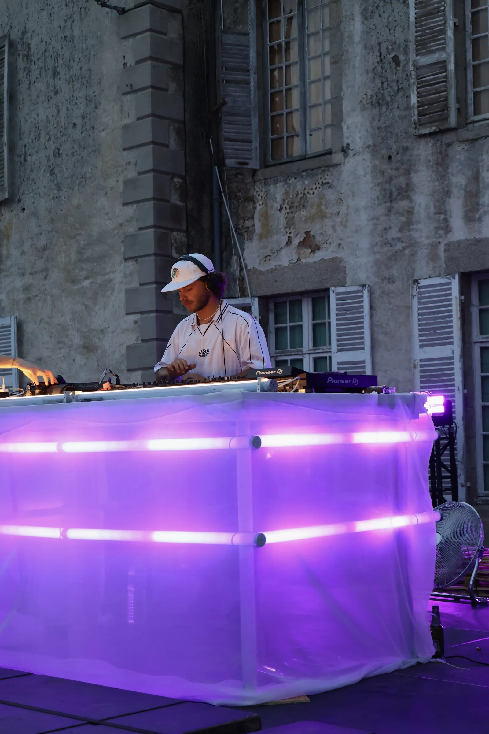 DJ mixant en soirée sur une scène éclairée en violet, devant la façade d'un bâtiment ancien.