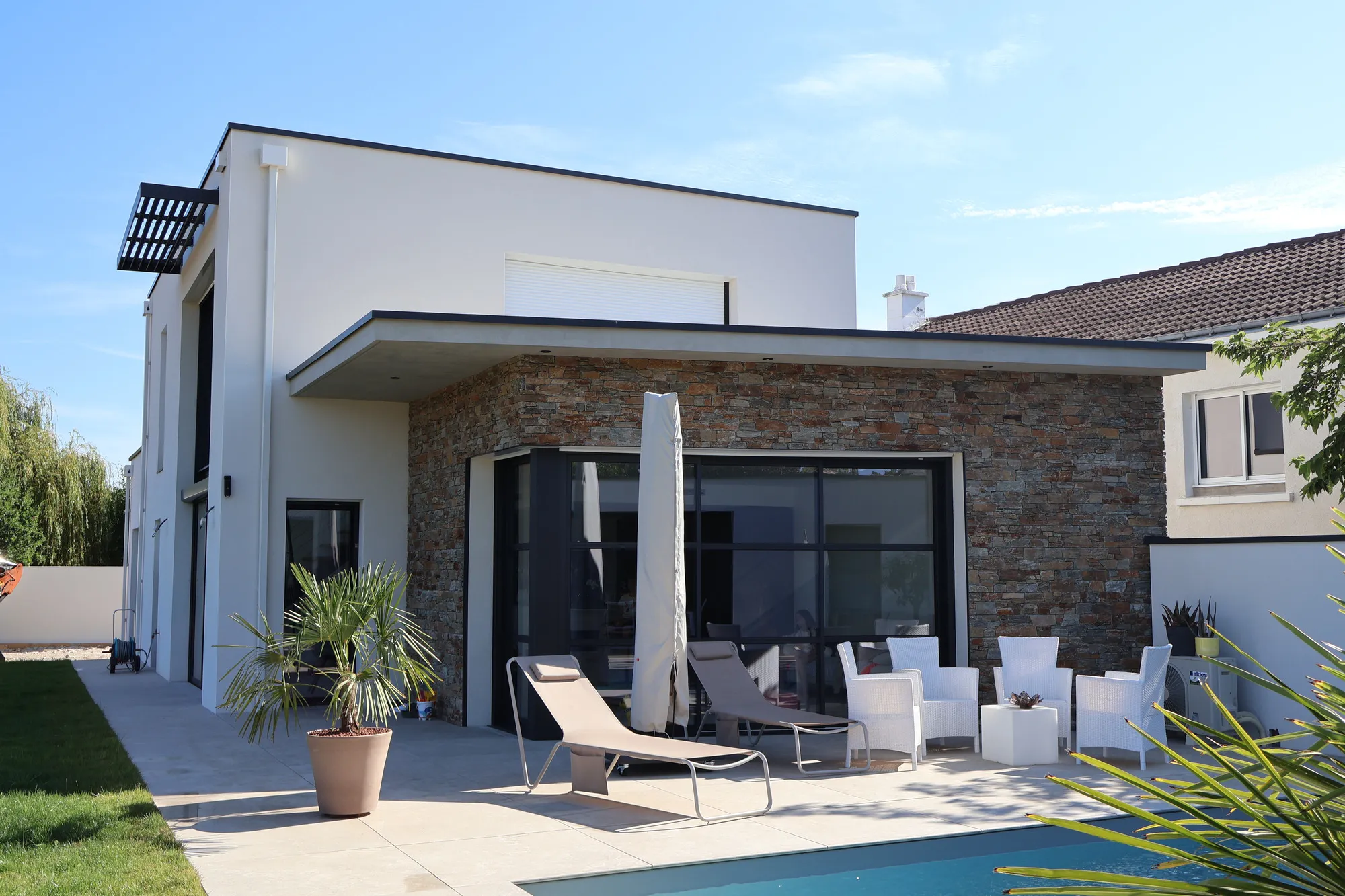 Façade jardin d'une maison moderne en pierre et enduit blanc, terrasse aménagée de mobilier en osier autour de la piscine.