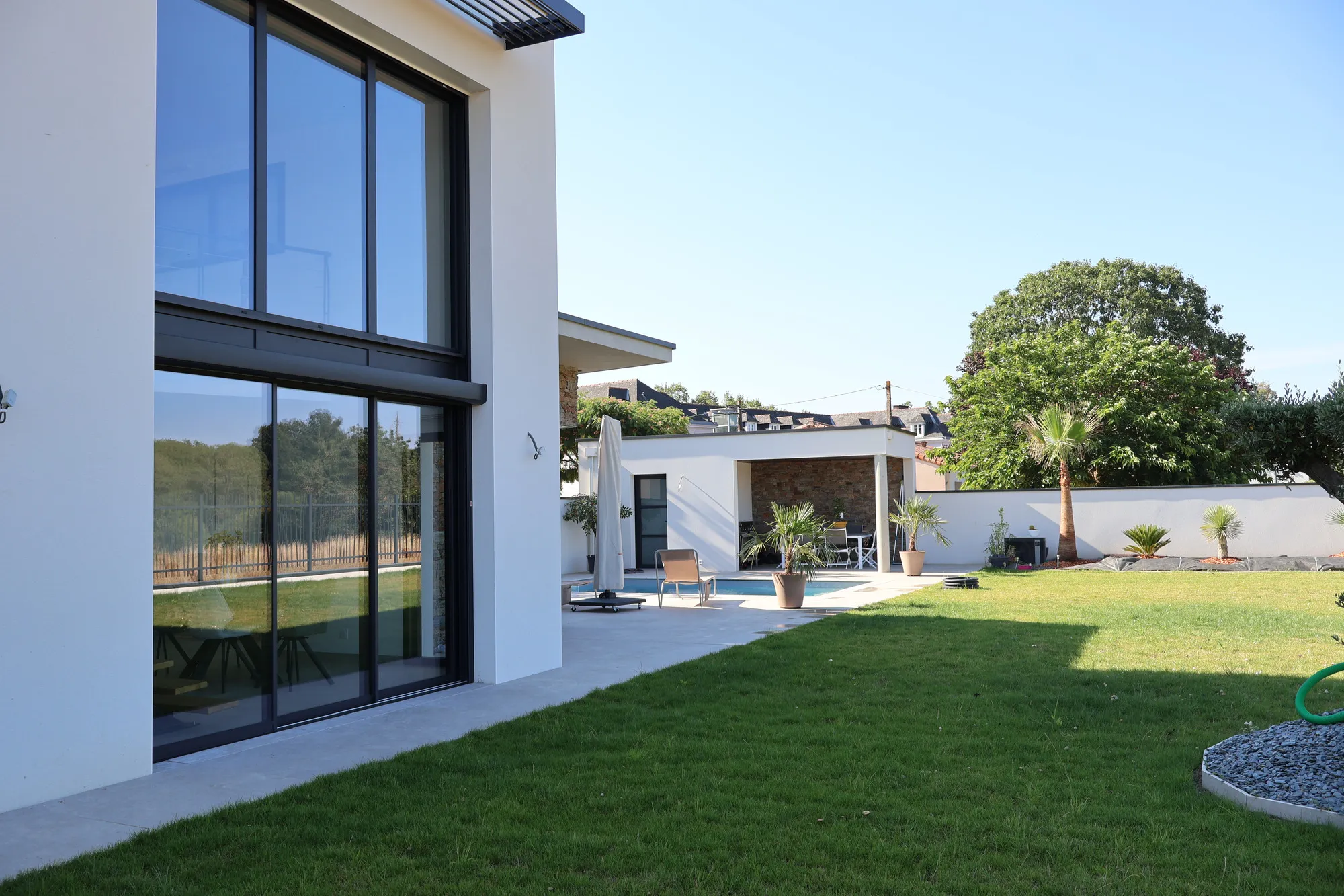 Façade arrière d'une maison C3P moderne et blanche, le jour de sa réception : larges baies vitrées, terrasse, piscine et jardin planté de palmiers en pot.