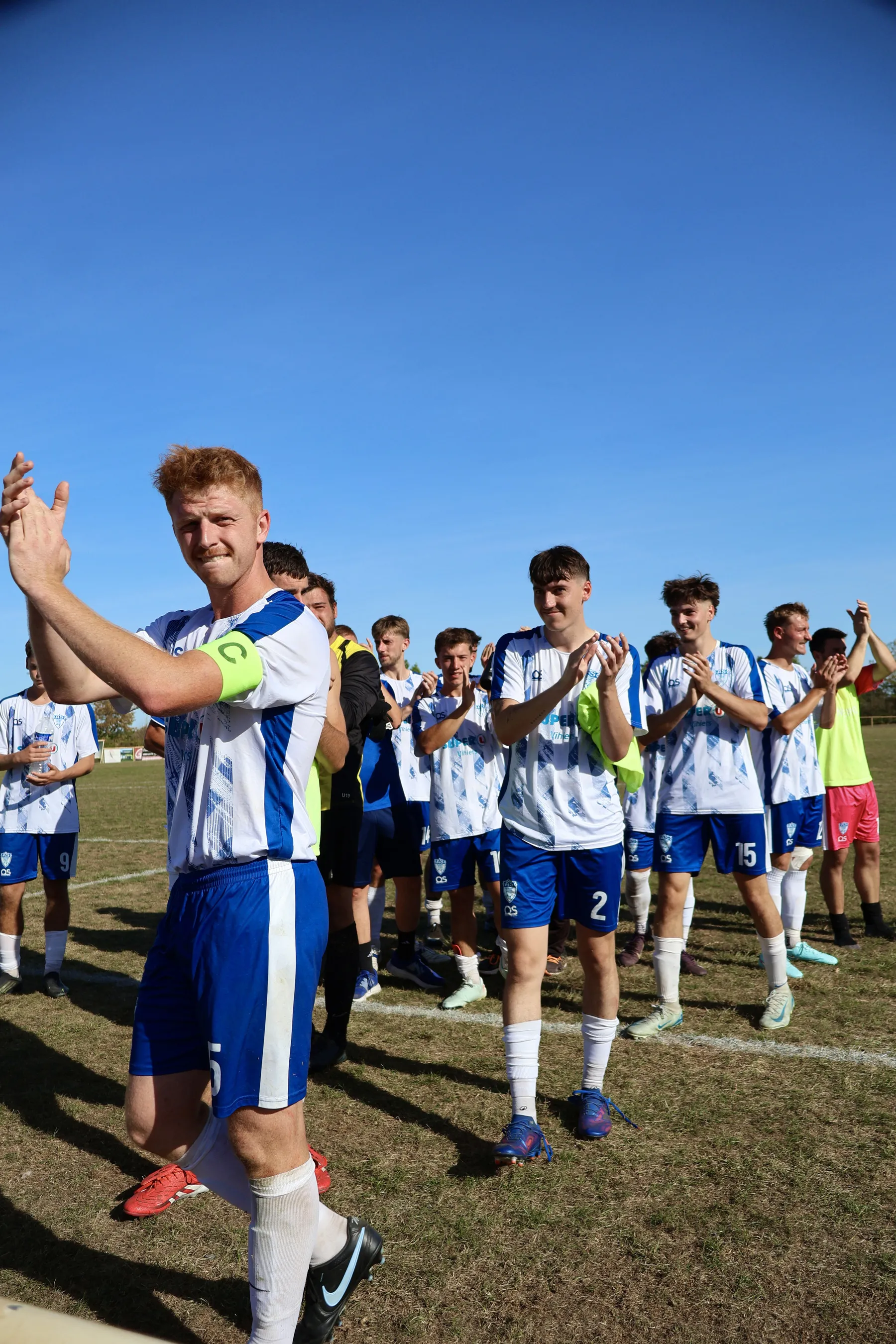 Le capitaine de l'ASSHVSP applaudit, devant ses coéquipiers en maillot blanc et bleu alignés sur le terrain.