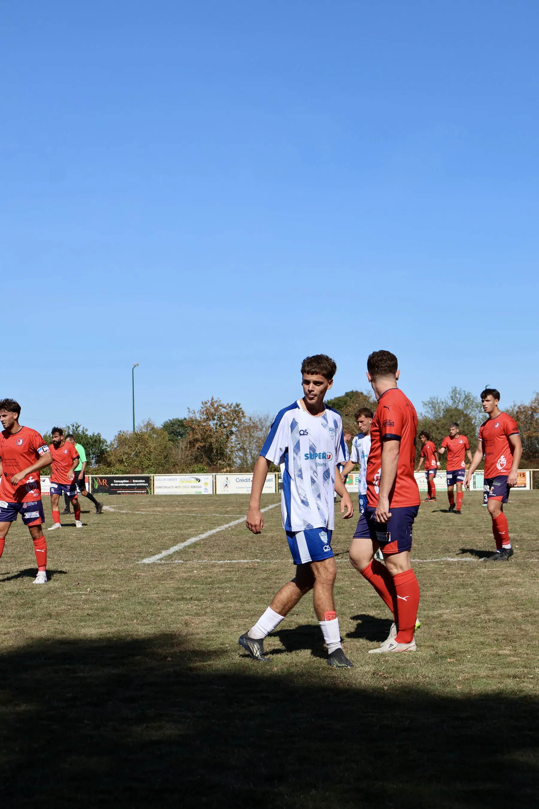 Duel au milieu du terrain entre un joueur en blanc et bleu et deux adversaires en rouge.